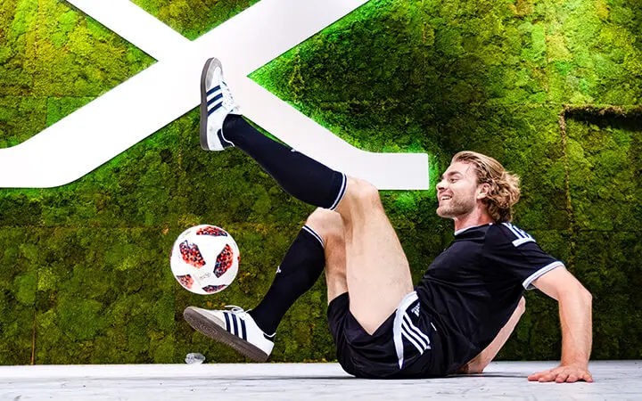 Ein Freestyle-Künstler in einem schwarzen Fußballtrikot und gestreiften Stutzen balanciert auf dem Boden und jongliert gekonnt einen Fußball mit dem Fuß vor einer grünen Mooswand mit einem großen weißen geometrischen Muster.
