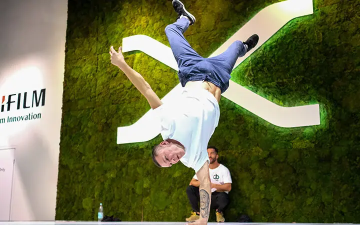 Ein Freestyle-Künstler macht einen einarmigen Handstand vor einer grünen Mooswand mit einem großen weißen X, während eine andere Person im Hintergrund auf dem Boden sitzt.