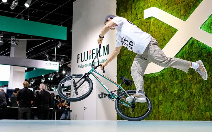 Ein Freestyle Künstler BMX-Fahrer führt in der Nähe eines Fujifilm-Standes einen Trick aus. Er balanciert auf einem Fuß, während mehrere Personen im Hintergrund zusehen.