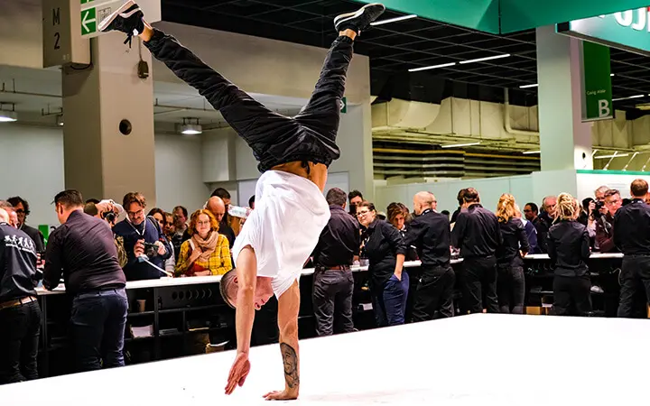 Ein Freestyle-Künstler führt auf der Bühne einen einhändigen Handstand vor, während eine Menschenmenge in einem Veranstaltungsraum zuschaut und Fotos macht.