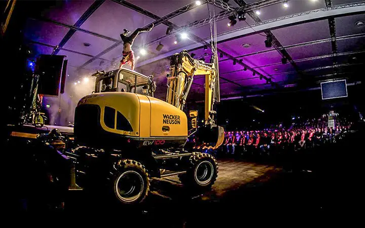 Ein Artist vollführt einen Handstand auf dem Arm eines Wacker Neuson Baggers während einer Urban Street Show, die mit dramatischer Beleuchtung und einem faszinierten Publikum in einer großen Halle inszeniert wird.