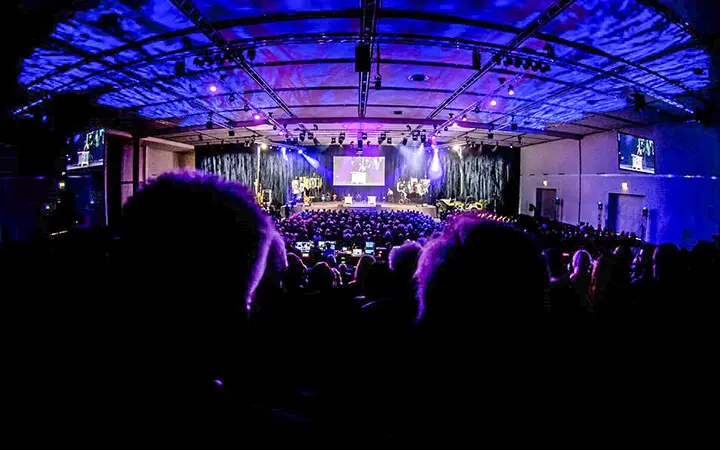 Ein großes Publikum sitzt in einem schwach beleuchteten Auditorium mit violetter und blauer Beleuchtung und blickt auf eine Bühne mit Bildschirmen, auf denen Text während einer Indoor-Veranstaltung oder Präsentation der Urban Street Show angezeigt wird.