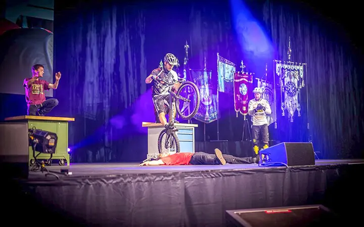 Ein Radfahrer mit Helm vollführt einen Stunt, indem er während einer Urban Street Show mit dem Fahrrad über eine auf der Bühne liegende Person springt, während zwei andere zuschauen; bunte Banner und dramatische Bühnenbeleuchtung bilden die Kulisse.
