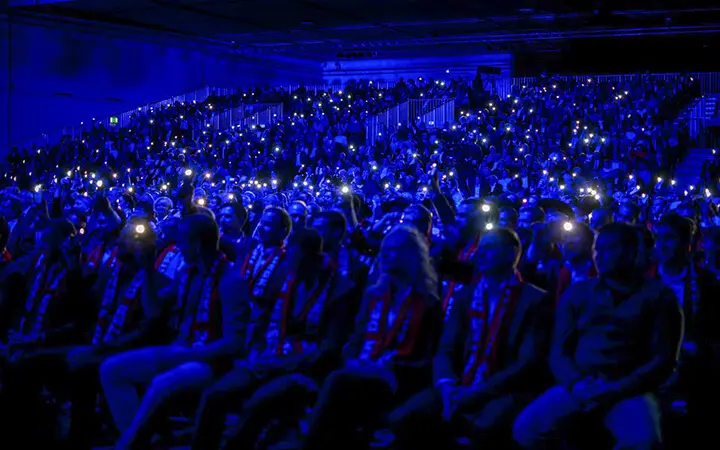 Ein großes Publikum sitzt in einem abgedunkelten, mit blauem Licht beleuchteten Saal, als ob es sich zu einer Urban Street Show versammelt hätte. Viele Menschen halten leuchtende Handys oder Taschenlampen in die Höhe und bilden ein Meer aus funkelnden Lichtern.