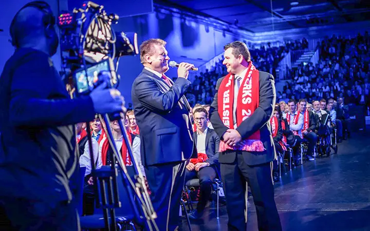 Zwei Männer in Anzügen und mit roten Schals sprechen auf der Bühne einer Indoor Urban Street Show, einer hält ein Mikrofon. Ein Kameramann filmt sie, während das Publikum im Hintergrund zusieht. Die Beleuchtung ist blau und lila.