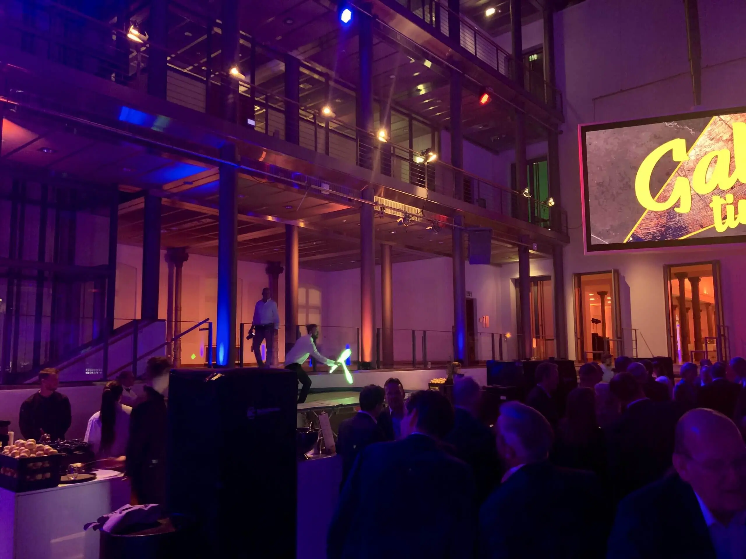 Eine Menschenmenge beobachtet eine Bühnenvorstellung in einem modernen, mehrstöckigen Veranstaltungsort mit bunter Beleuchtung. Ein Freestyle-Künstler jongliert auf der Bühne mit leuchtenden Objekten, während auf einer großen Leinwand das Wort "Gala" erscheint.