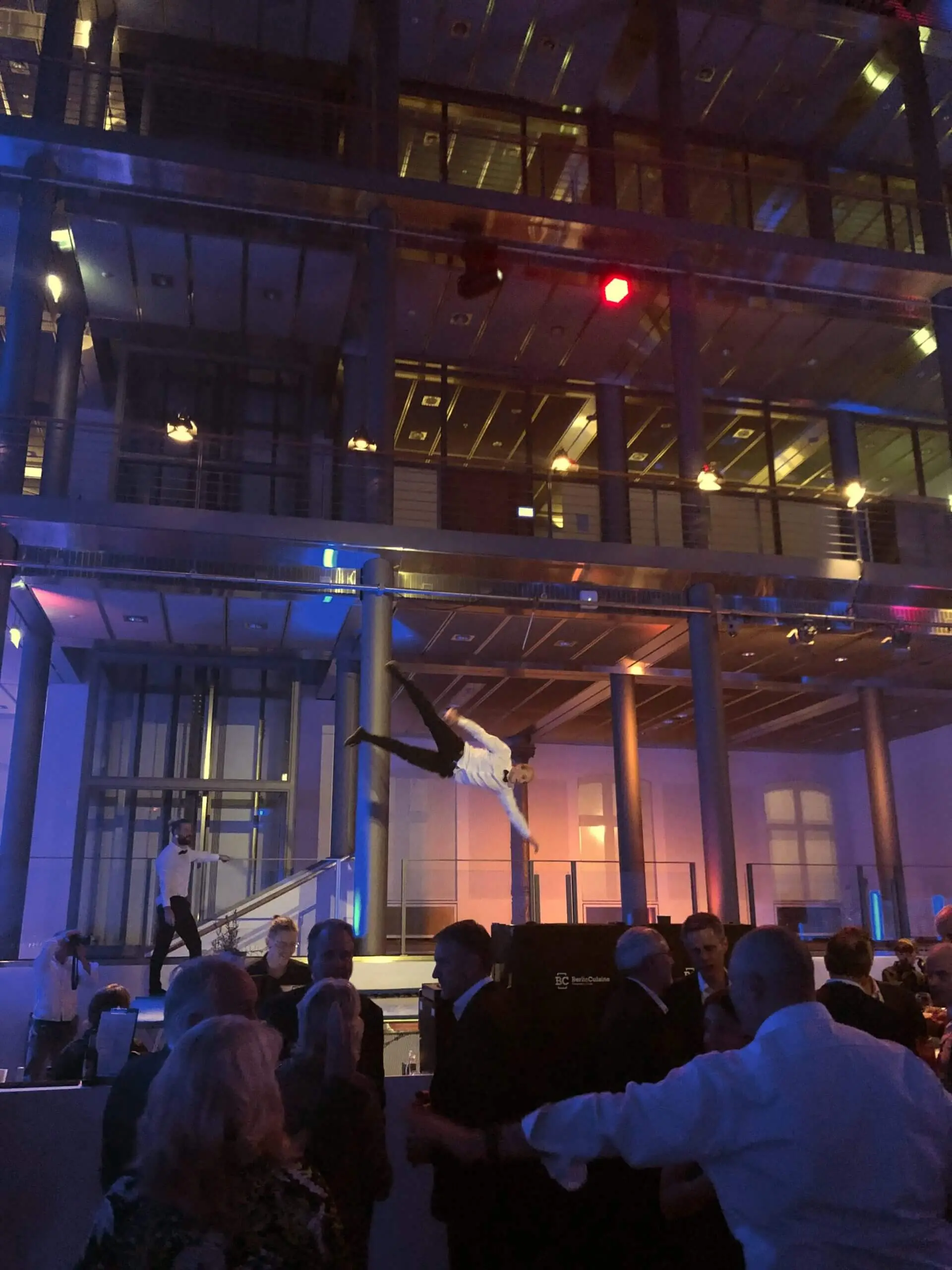 Ein Freestyle-Künstler vollführt in der Luft einen akrobatischen Salto über einer Menschenmenge bei einer Indoor-Veranstaltung mit moderner Glasarchitektur, dramatischer Beleuchtung und Zuschauern unter ihm.