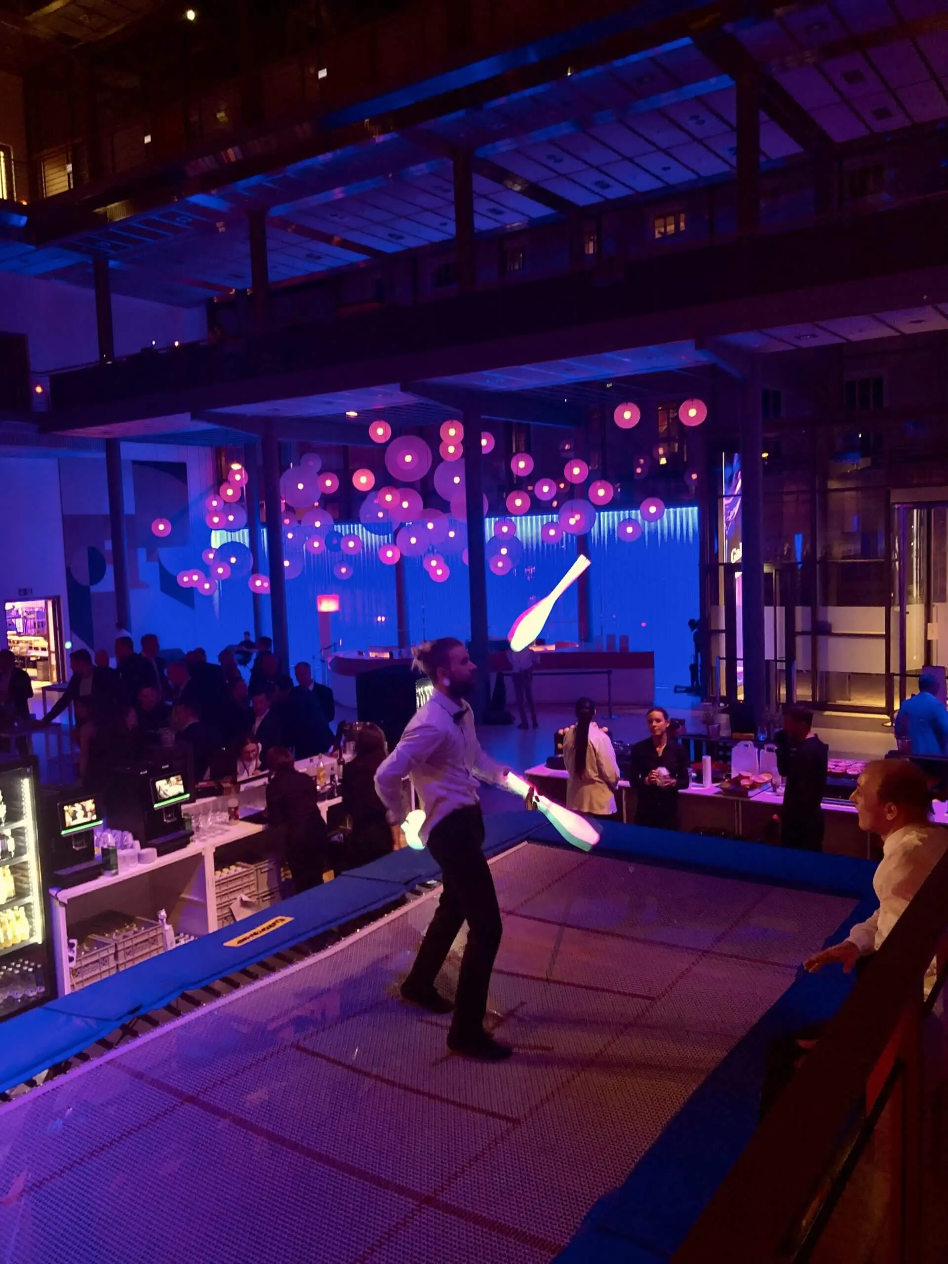 Ein Freestyle-Künstler jongliert mit Stiften, während er auf einem Trampolin bei einer lebhaften Indoor-Veranstaltung steht, mit Menschen, die sich unterhalten, einem Barbereich und lila Hängelampen im Hintergrund.
