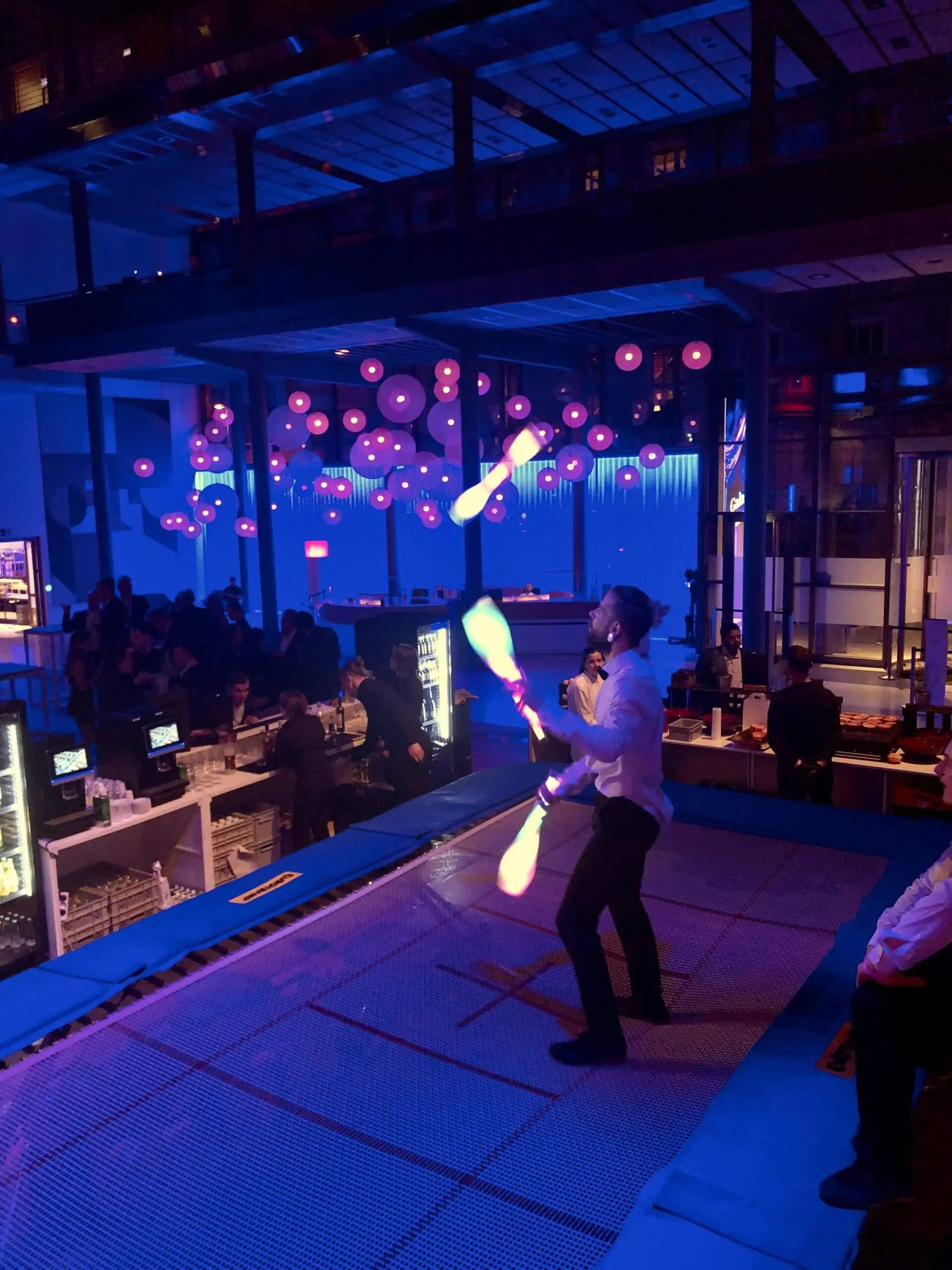 Ein Freestyle-Künstler im weißen Hemd jongliert mit leuchtenden Stiften auf einem Trampolin bei einer lebhaften Indoor-Veranstaltung mit bunten Lichtern und hängenden Dekorationen, während im Hintergrund Menschen zusehen und sich unterhalten.