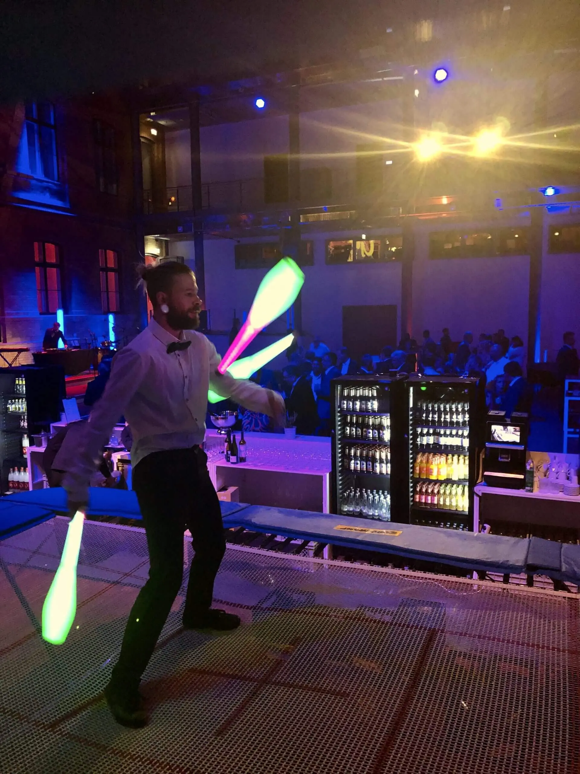 Ein Mann in weißem Hemd und Fliege, der als Freestyle-Künstler auftritt, jongliert drei leuchtende Keulen bei einer lebhaften Indoor-Veranstaltung mit bunter Beleuchtung und einem sitzenden Publikum im Hintergrund.