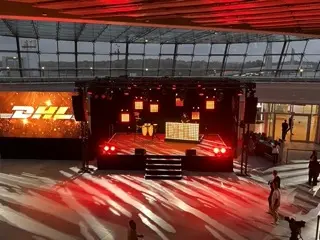Im Innenraum ist eine rot beleuchtete Bühne aufgebaut, auf der Musikinstrumente und Leuchttafeln für eine LED-Trommelshow zu sehen sind. Auf der linken Seite wird auf einem großen Bildschirm das DHL-Logo angezeigt. Ein paar Menschen sind auf der Freifläche verstreut.
