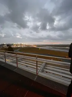 Blick auf die Start- und Landebahn eines Flughafens in der Abenddämmerung, mit Lichtern, die die Start- und Landebahn und die Terminalgebäude in der Ferne beleuchten. Der Himmel ist wolkenverhangen, und die Bänke auf einer Aussichtsplattform erwecken Vorfreude - wie das Warten auf den Beginn einer LED-Trommel-Show.