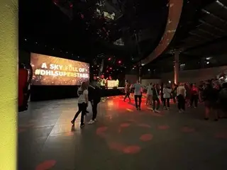 Eine Gruppe von Menschen versammelt sich in einem schwach beleuchteten Veranstaltungsraum, während eine LED-Trommel-Show den Raum in Schwung bringt. Auf einer großen Leinwand ist in weißer Schrift A SKY FULL OF #DHLSUPERSTARS zu sehen, während auf dem Boden orangefarbene Lichteffekte zu sehen sind.