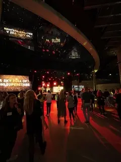 Die Menschen versammeln sich in einem schwach beleuchteten Veranstaltungsraum mit roter Beleuchtung, großen Bildschirmen, die Texte anzeigen, und einer Bühne im Hintergrund. Die lebhafte Atmosphäre deutet auf eine elektrisierende Nacht hin, möglicherweise mit einer LED-Trommel-Show.