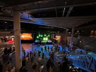 Eine nächtliche Veranstaltung im Freien, bei der sich die Menschen vor einer beleuchteten Bühne mit grünen und gelben Lichtern unter einer modernen Struktur mit Balken und Säulen versammeln. Die lebhafte Szene wird durch die farbenfrohe Beleuchtung einer elektrisierenden LED-Trommel-Show erhellt.