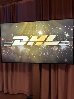 Eine große Leinwand zeigt das DHL-Logo vor einem funkelnden, beleuchteten Hintergrund, der eine lebendige Atmosphäre schafft, die an eine LED-Trommel-Show erinnert. Die Leinwand ist vor beigefarbenen Vorhängen angebracht.