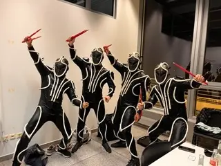 Vier Personen in passenden schwarzen Bodysuits mit leuchtenden weißen Linien und Vollhelmen posieren dynamisch in Innenräumen, wobei jede von ihnen rote Trommelstöcke hält und eine beeindruckende LED-Trommelshow mit einem futuristischen Sci-Fi-Thema kreiert.