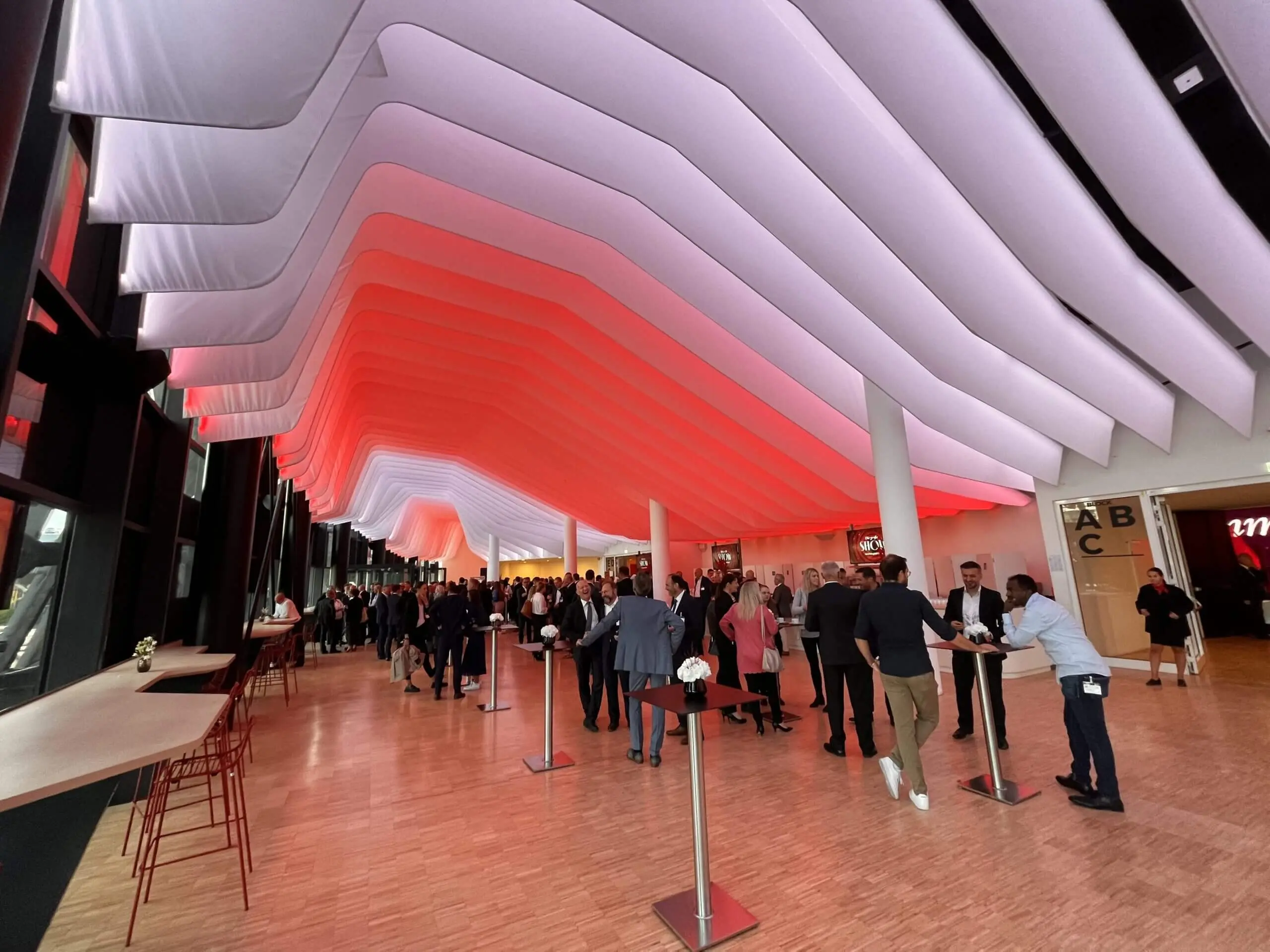 In einem modernen Veranstaltungsraum mit einer dramatisch gewellten Decke, die in Rot und Weiß beleuchtet ist, trifft sich eine große Menschenmenge an hohen Tischen und unterhält sich. Hohe Fenster säumen eine Seite, und auf dem Holzboden sind rote Stühle gestapelt, die den Rahmen für eine aufregende LED-Trommel-Show bilden.