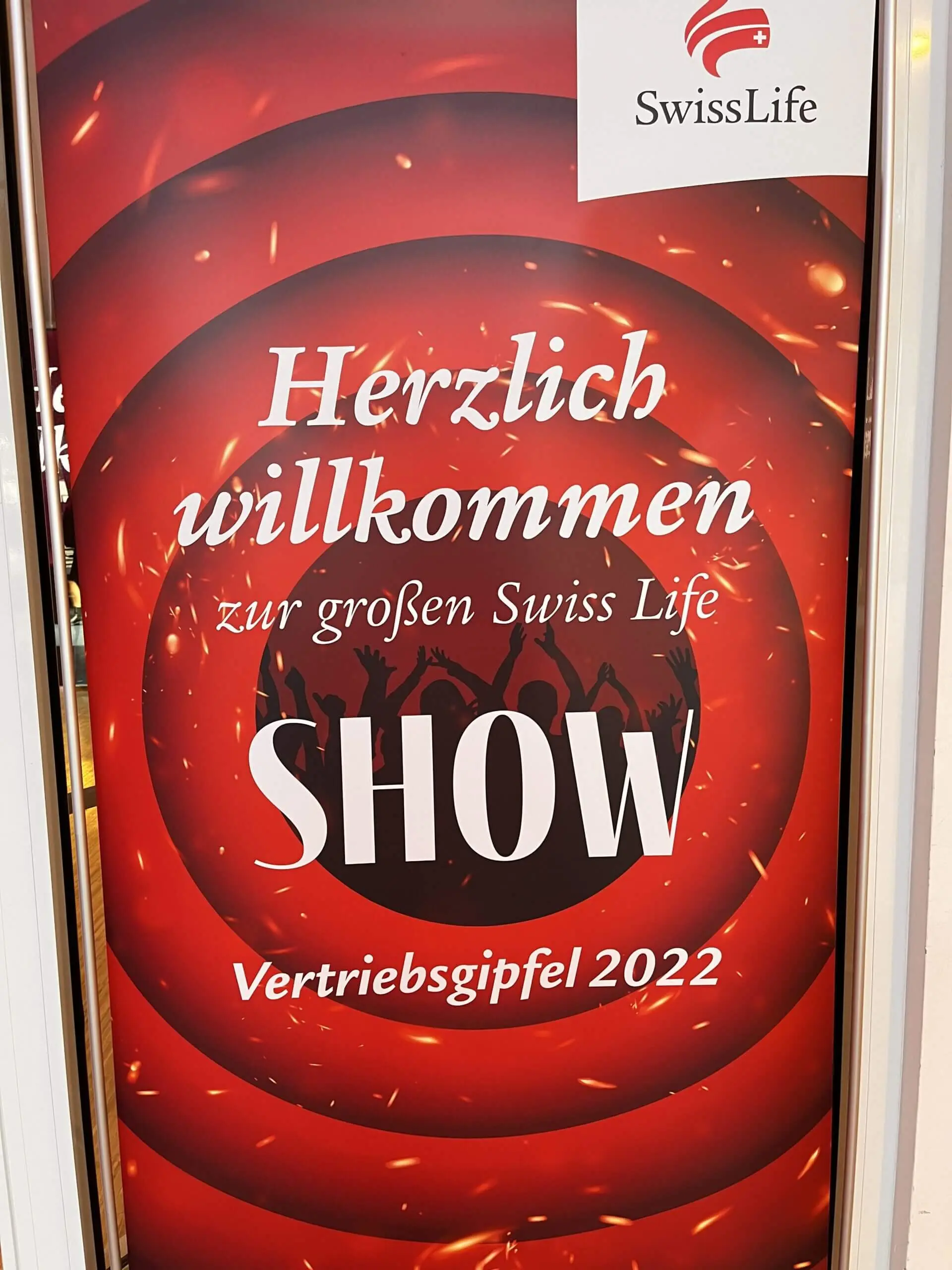 Rot-schwarzes Plakat mit dem Swiss Life-Logo und deutschem Text: Herzlich willkommen zum grossen Swiss Life SHOW Vertriebsgipfel 2022. Silhouetten von jubelnden Menschen und ein Hinweis auf eine elektrisierende LED-Trommel-Show im Hintergrund.