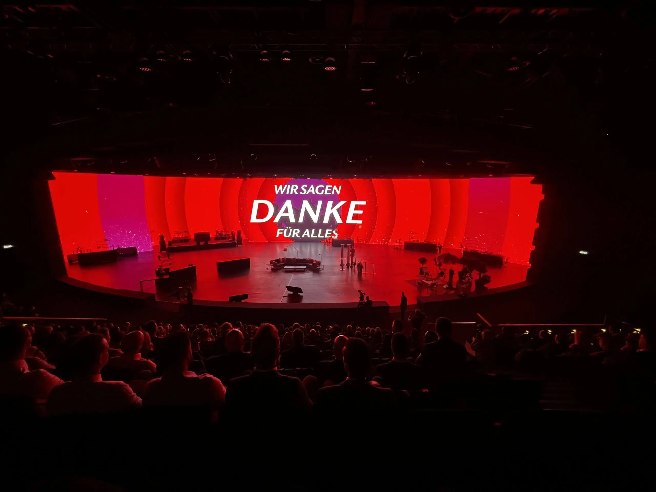 Eine große Bühne mit roter Beleuchtung und einer gebogenen Leinwand mit der Aufschrift "WIR SAGEN DANKE FÜR ALLES" ist auf das Publikum ausgerichtet. Auf der Bühne befinden sich mehrere Personen und Musikinstrumente, darunter ein LED-Drum-Show-Setup.