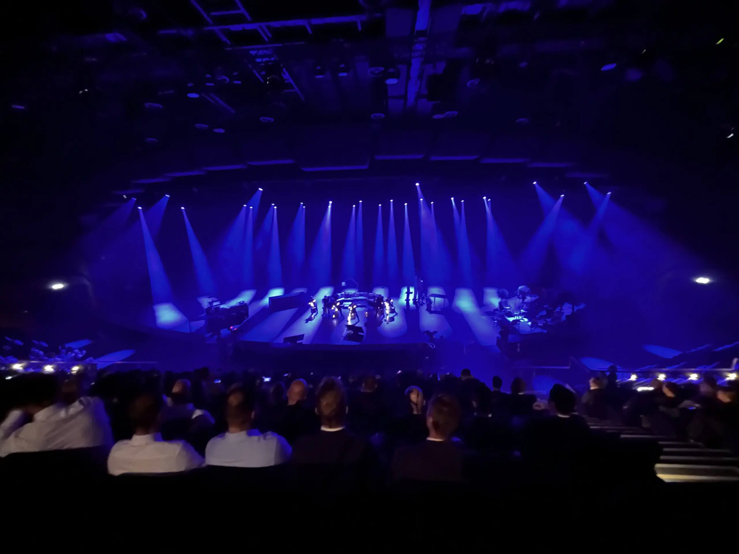Eine Bühnenaufführung, die vom Publikum aus gesehen wird, mit blauen Scheinwerfern und einer LED-Trommel-Show, die Musiker und Darsteller auf der Bühne beleuchtet, während das sitzende Publikum in einem dunklen Theater zusieht.