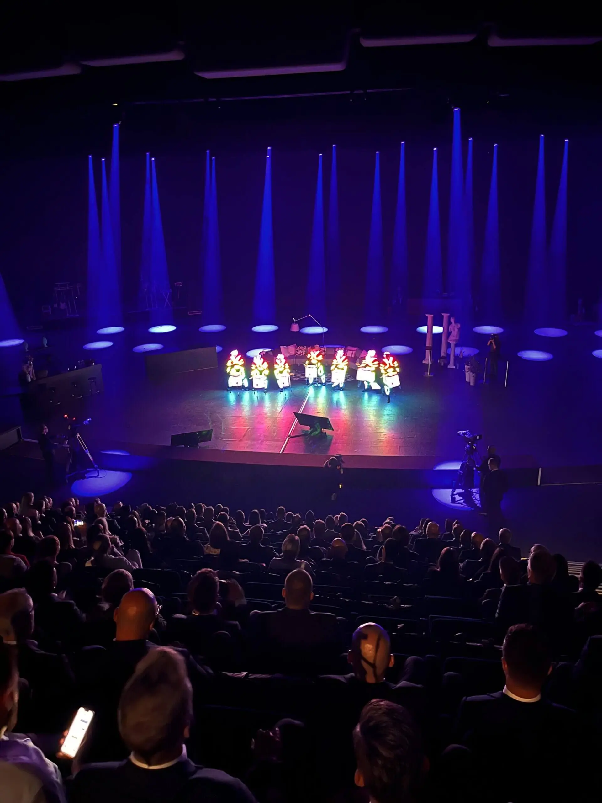 Ein Theaterpublikum beobachtet eine Gruppe von Darstellern in farbenfrohen Kostümen und hellen Scheinwerfern, während blaues und violettes Licht die Stimmung für eine elektrisierende LED-Trommel-Show auf der Bühne erzeugt.