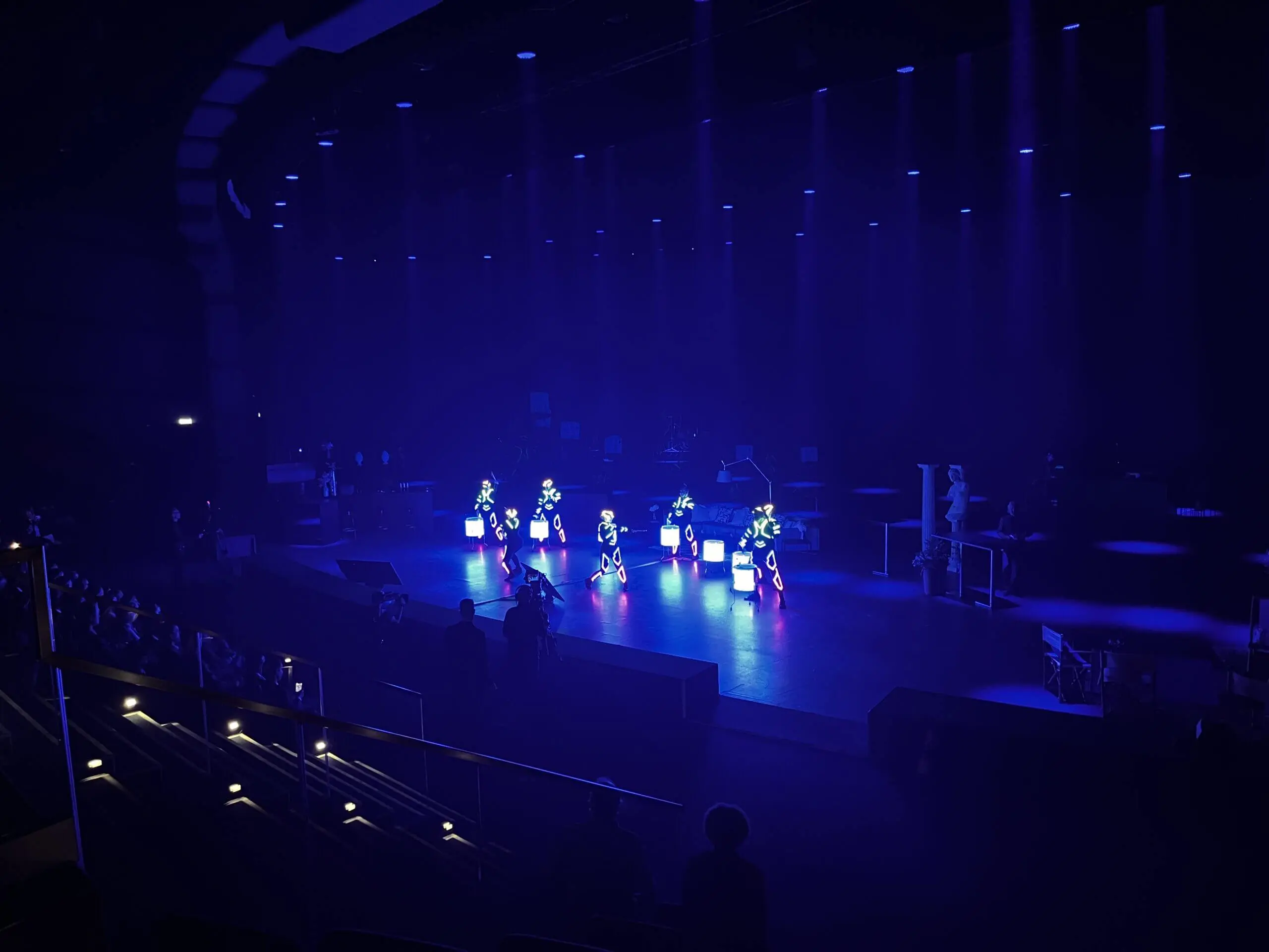 Eine dunkle Bühne wird mit blauen und violetten Lichtern beleuchtet. LED-Drum-Show-Darsteller in beleuchteten Kostümen stehen in Formation vor der Bühne, während Silhouetten von Menschen aus dem Publikum und von der Bühnenseite aus zusehen.