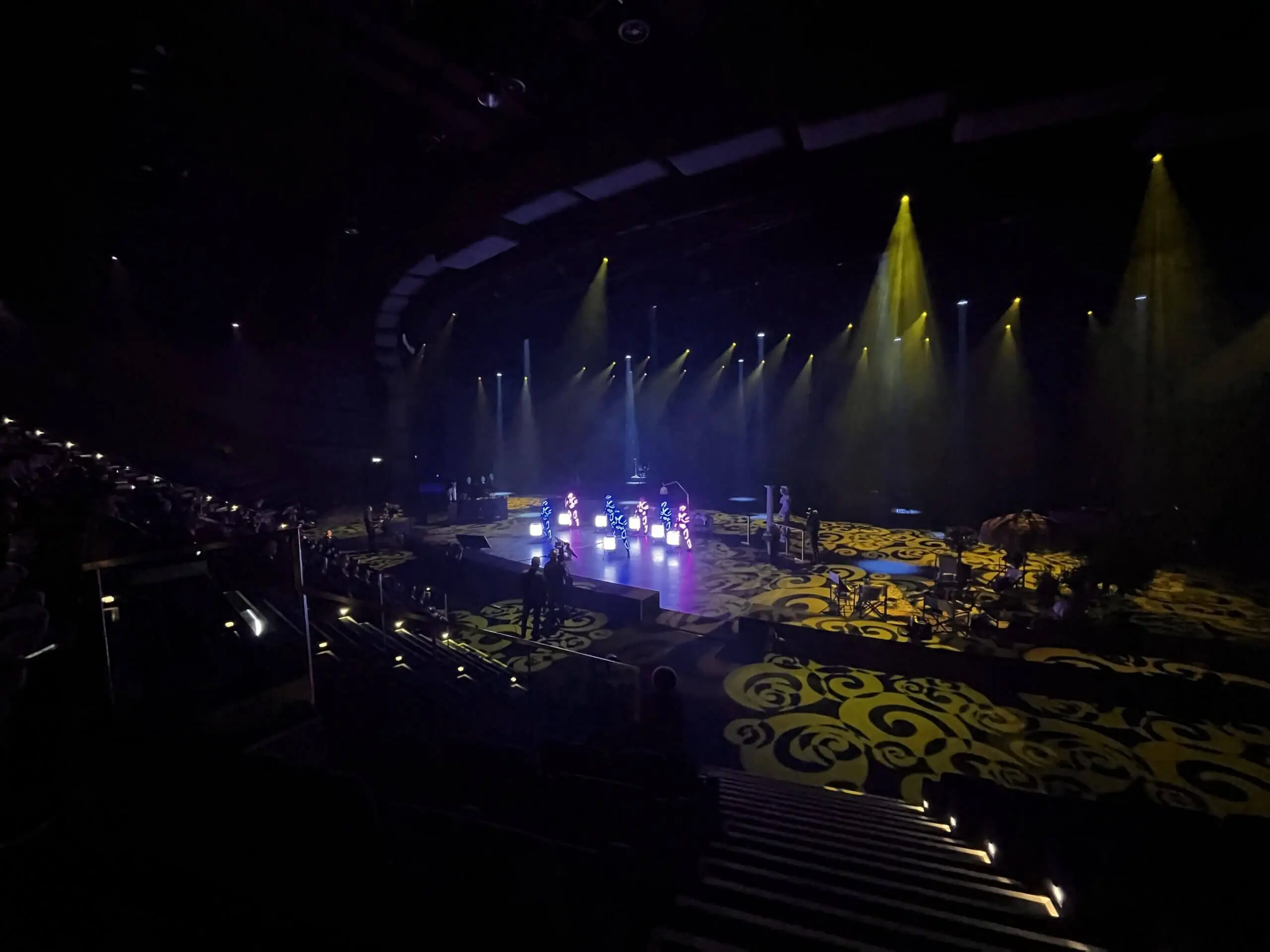 Ein dunkles Theater mit gelben Bühnenscheinwerfern, die verschlungene Muster auf dem Boden beleuchten. Die Darsteller einer LED-Trommel-Show sind auf der Bühne zu sehen, während leere Sitze den Vordergrund füllen und Zuschauer an den Seiten zu sehen sind.