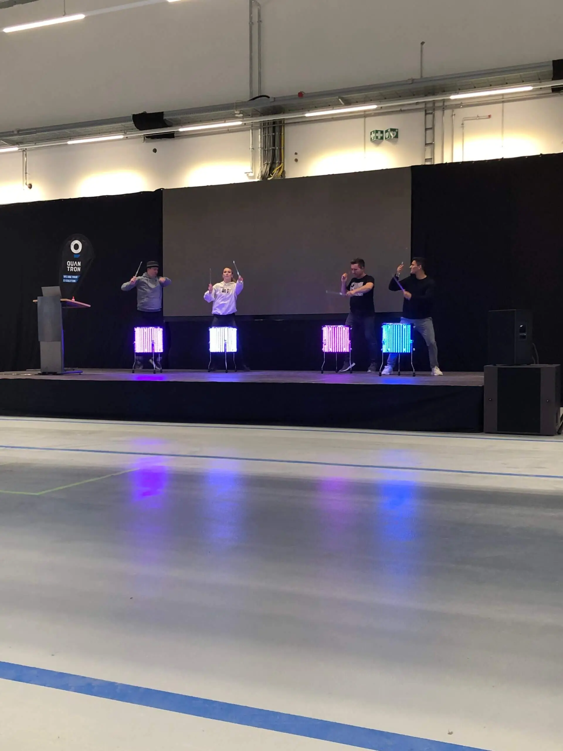 Vier Personen stehen auf einer Bühne, jede hinter einem farbenfrohen LED-Drumshow-Setup, und spielen mit erhobenen Trommelstöcken. Der schwarze Hintergrund und der reflektierende Boden verstärken die leuchtenden Lichter der beleuchteten Trommeln.
