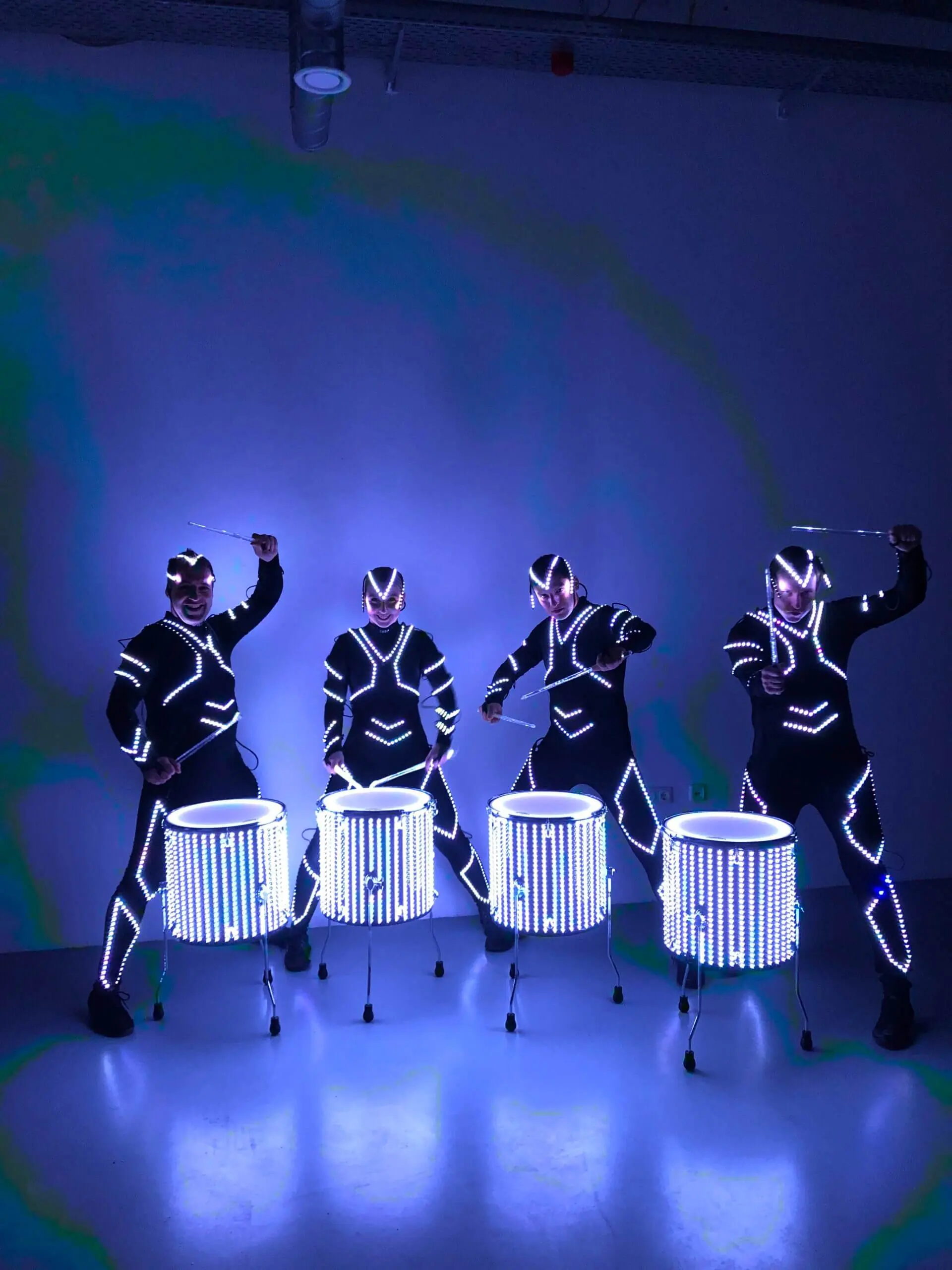 Vier Darsteller in leuchtenden LED-Anzügen liefern eine elektrisierende LED-Trommel-Show, indem sie in einem dunklen Raum auf beleuchteten Trommeln spielen. Helles blaues und weißes Licht beleuchtet ihre Figuren und Instrumente und schafft eine lebendige, futuristische Szene.