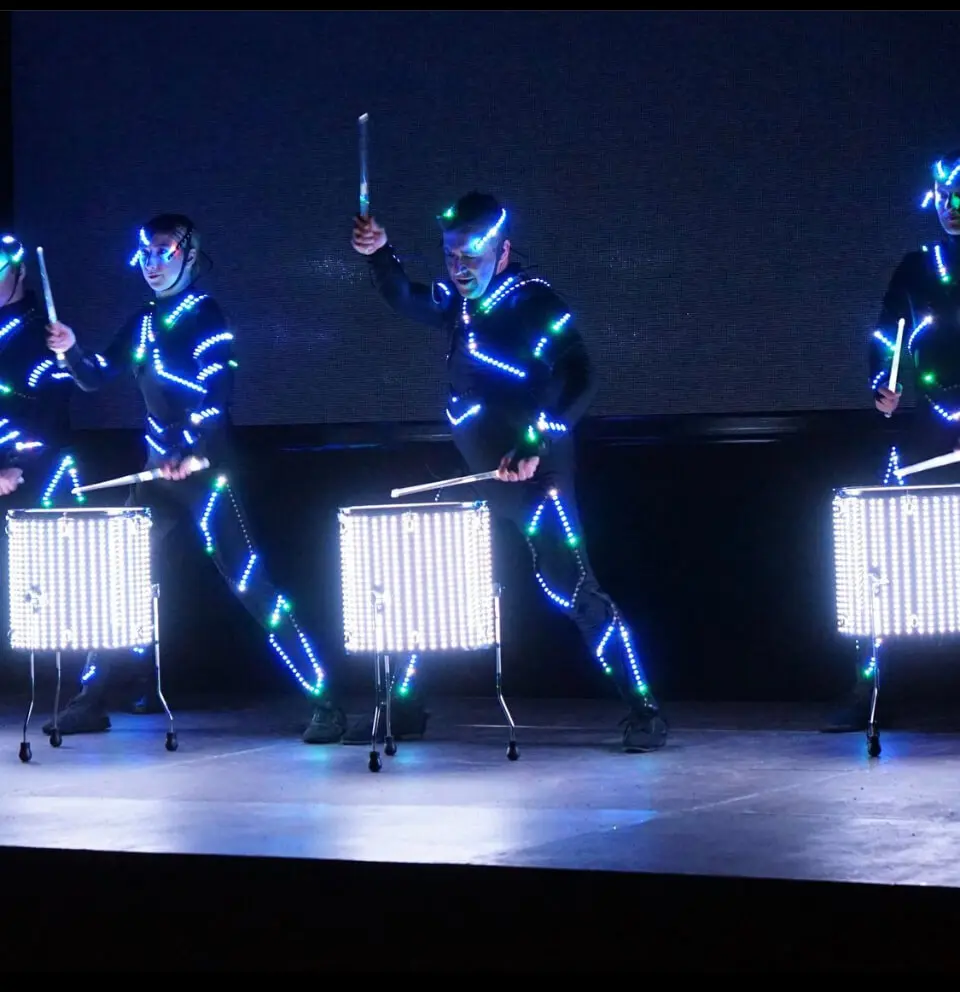 Vier Schlagzeuger in schwarzen Anzügen mit blauen LED-Lichtern führen eine energiegeladene LED-Trommelshow auf. Sie spielen auf beleuchteten Trommeln, die mit vertikalen Lichtmustern vor einem dunklen Hintergrund leuchten.