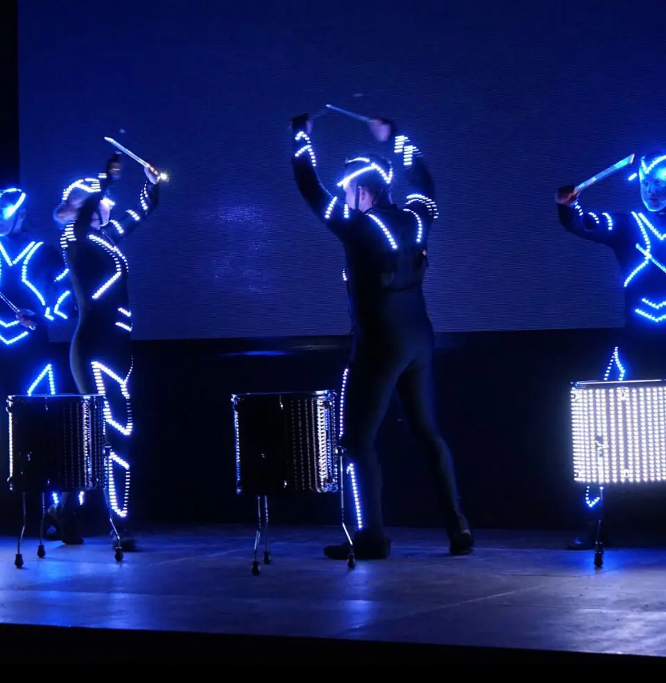 Drei Performer in schwarzen Anzügen mit blauen LEDs liefern eine elektrisierende LED-Trommel-Show, bei der sie auf einer dunklen Bühne mit blau beleuchtetem Hintergrund ein beleuchtetes Schlagzeug spielen. Ihre synchronisierten Bewegungen schaffen eine dynamische, futuristische Atmosphäre.