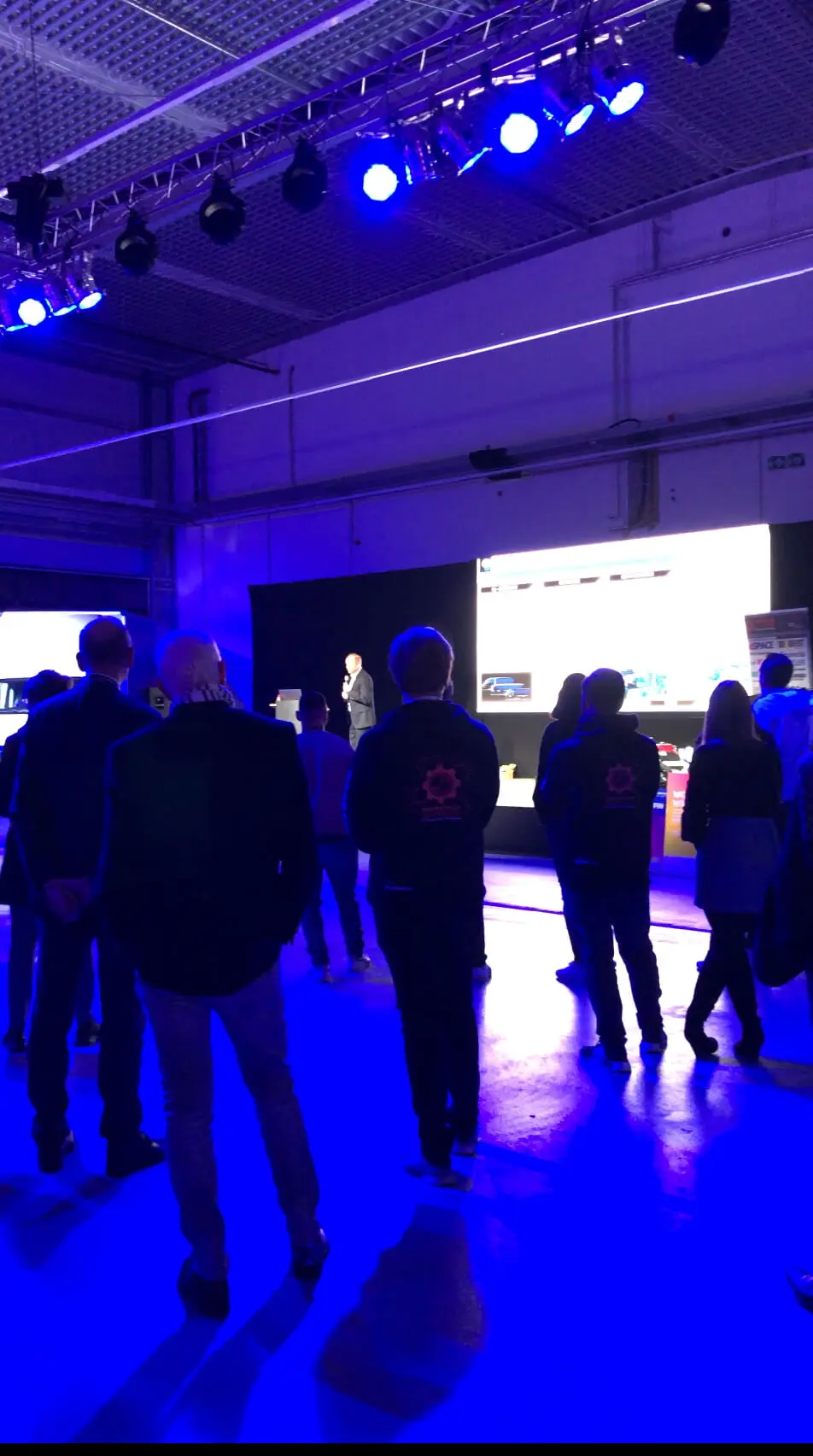 Eine Gruppe von Menschen steht in einem schwach beleuchteten, blau beleuchteten Raum und sieht sich eine Präsentation an, die auf eine große Leinwand projiziert wird, während ein LED-Drum-Show-Künstler die Bühne betritt und das Publikum mit leuchtenden Lichtern und Rhythmen in seinen Bann zieht.