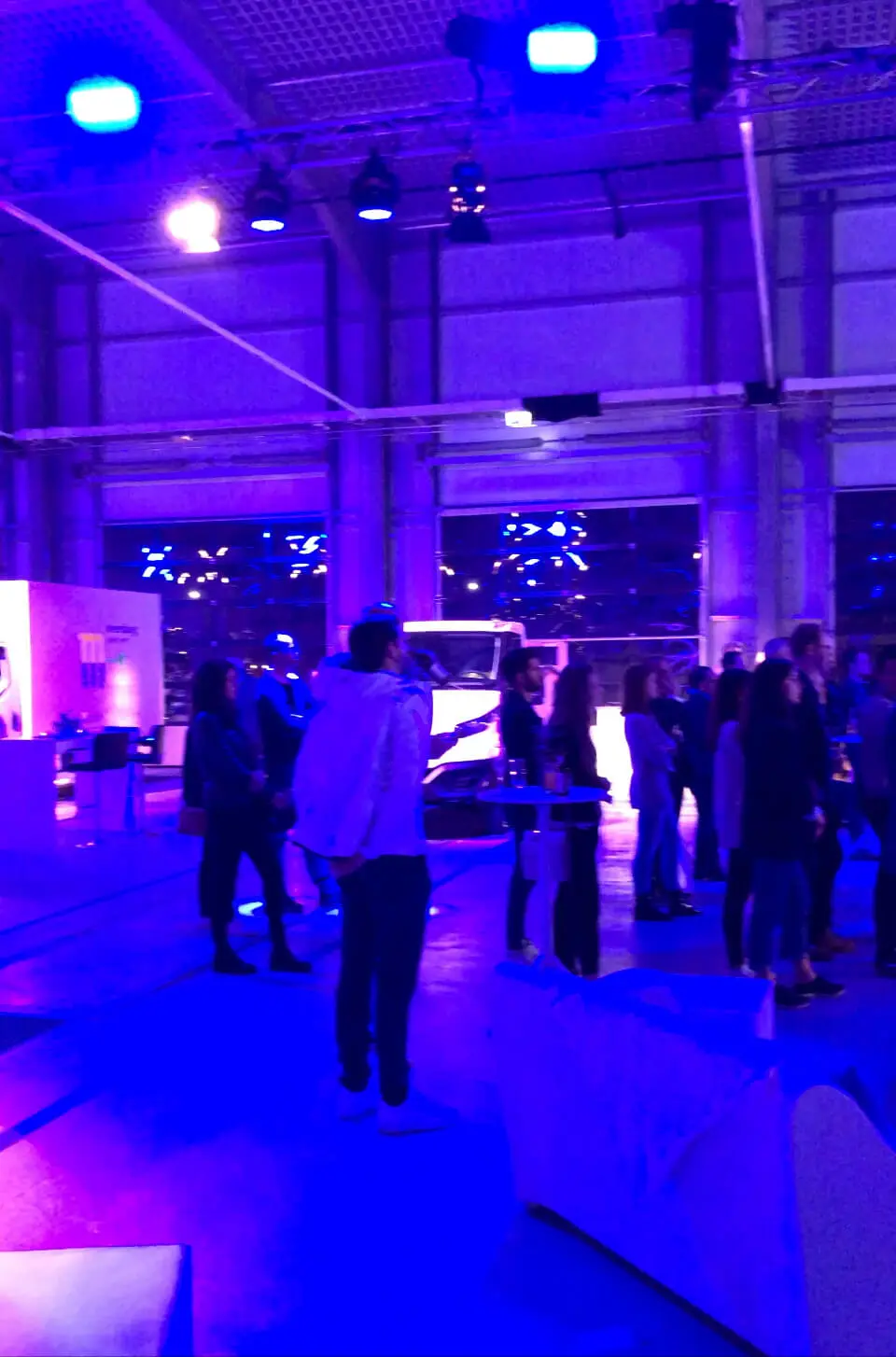 Eine Gruppe von Menschen versammelt sich in einem geräumigen, modernen Innenraum mit violetter und blauer Beleuchtung, LED-Trommelvorführungen, beleuchteten Displays und großen Fenstern im Hintergrund und knüpft Kontakte.