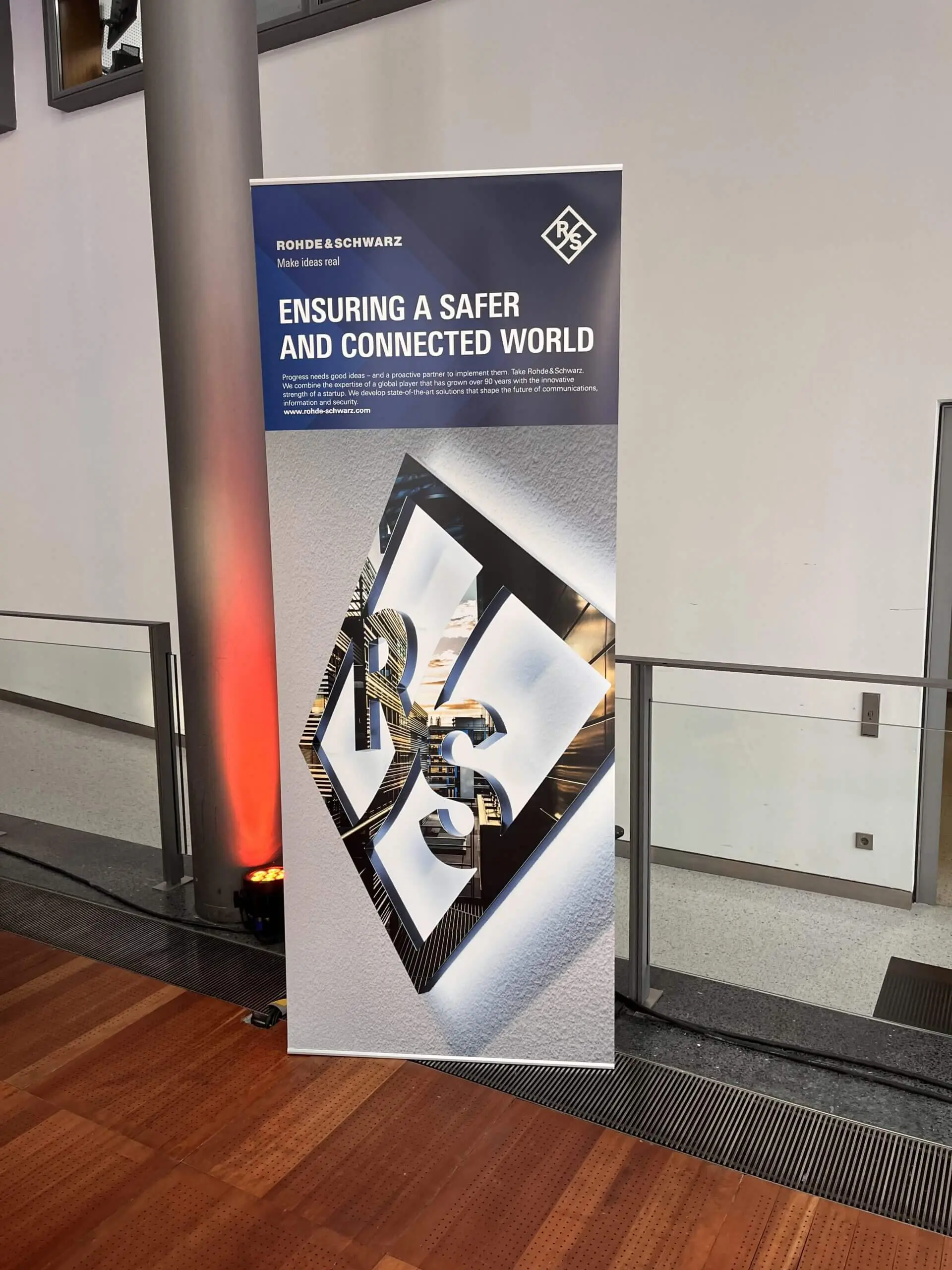 Ein stehendes Banner zeigt das Rohde & Schwarz-Logo und den Slogan Ensuring a Safer and Connected World, eingebettet in einen modernen Innenraum mit Holzfußboden - eine einladende Kulisse für jede Freestyle Künstler Performance.