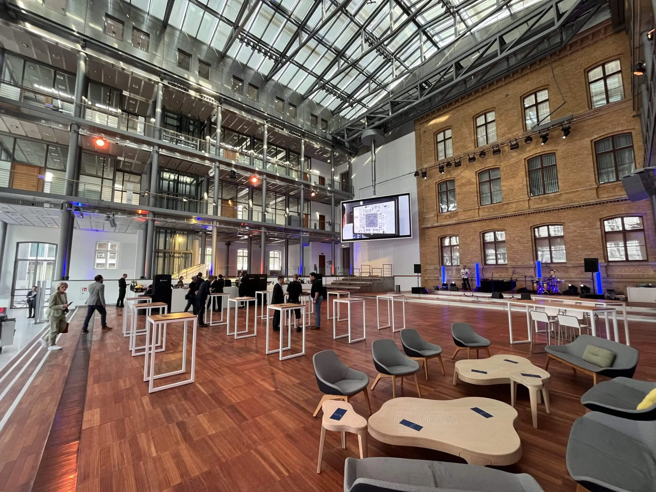 Ein geräumiger, moderner Veranstaltungsort mit Holzböden, hohen Glasdecken und sichtbaren Backsteinwänden. Stühle und Tische sind überall aufgestellt, während sich die Freestyle-Künstler vorbereiten, und vorne wird auf einer großen Leinwand eine Präsentation gezeigt. Die Leute bauen auf.