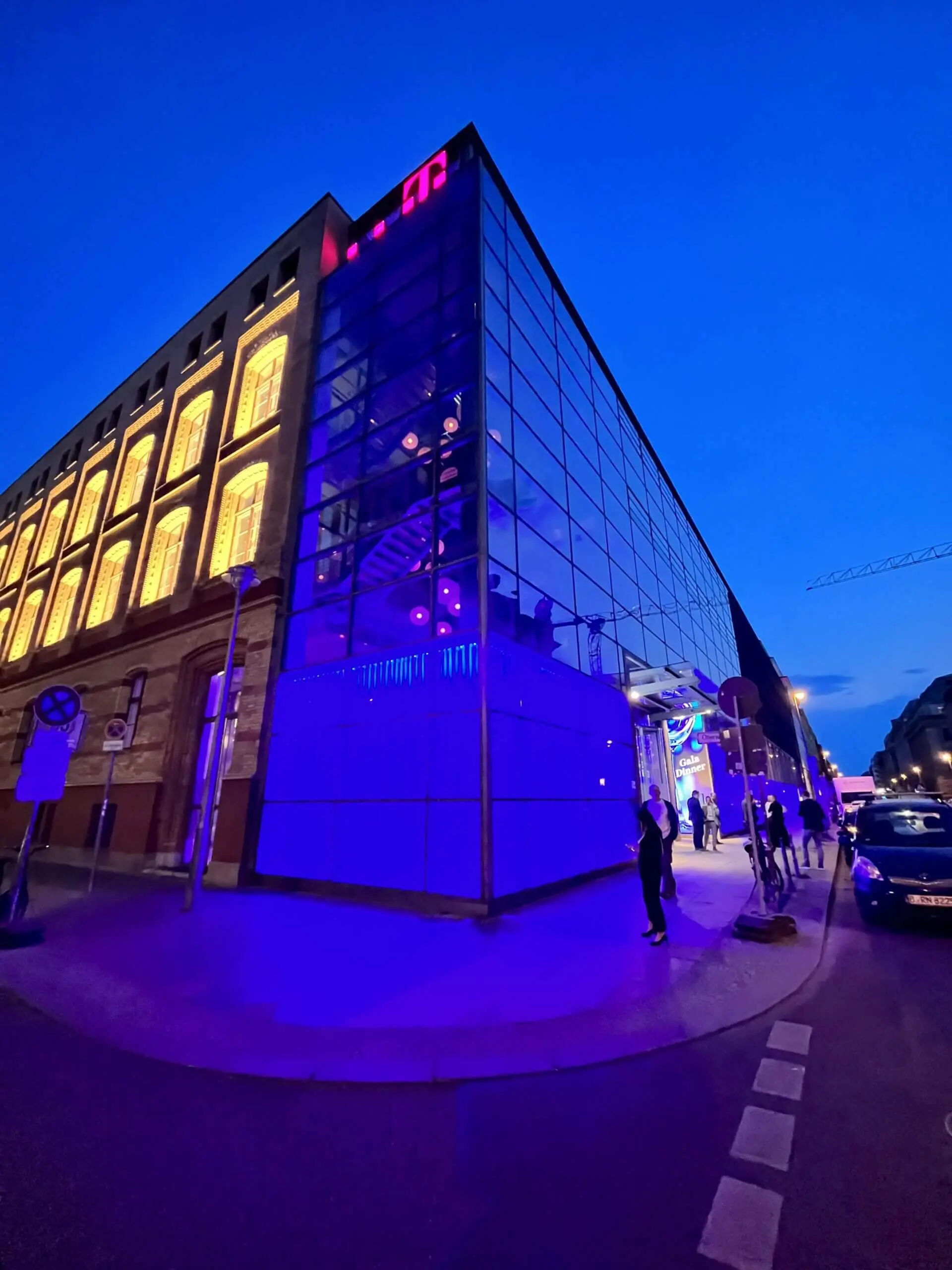 Ein modernes Glasgebäude mit lila und blauer Beleuchtung steht in der Abenddämmerung an einer Straßenecke. In der Nähe treten Freestyle-Künstler auf, während sich Menschen versammeln und ein Teil einer älteren Backsteinfassade mit gelben Lichtern beleuchtet wird.