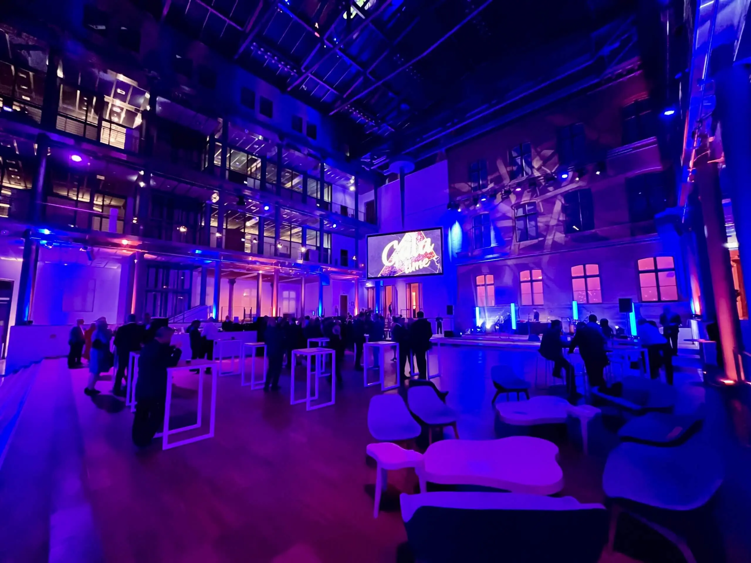 Ein geräumiger Veranstaltungsort im Innenbereich mit modernem Dekor und lila-blauer Beleuchtung. Die Leute sitzen an Stehtischen, während ein Freestyle-Künstler auftritt, während auf einem großen Bildschirm in leuchtenden Buchstaben Creative angezeigt wird. Die Decke ist mit Glaspaneelen versehen und die Atmosphäre ist lebhaft.