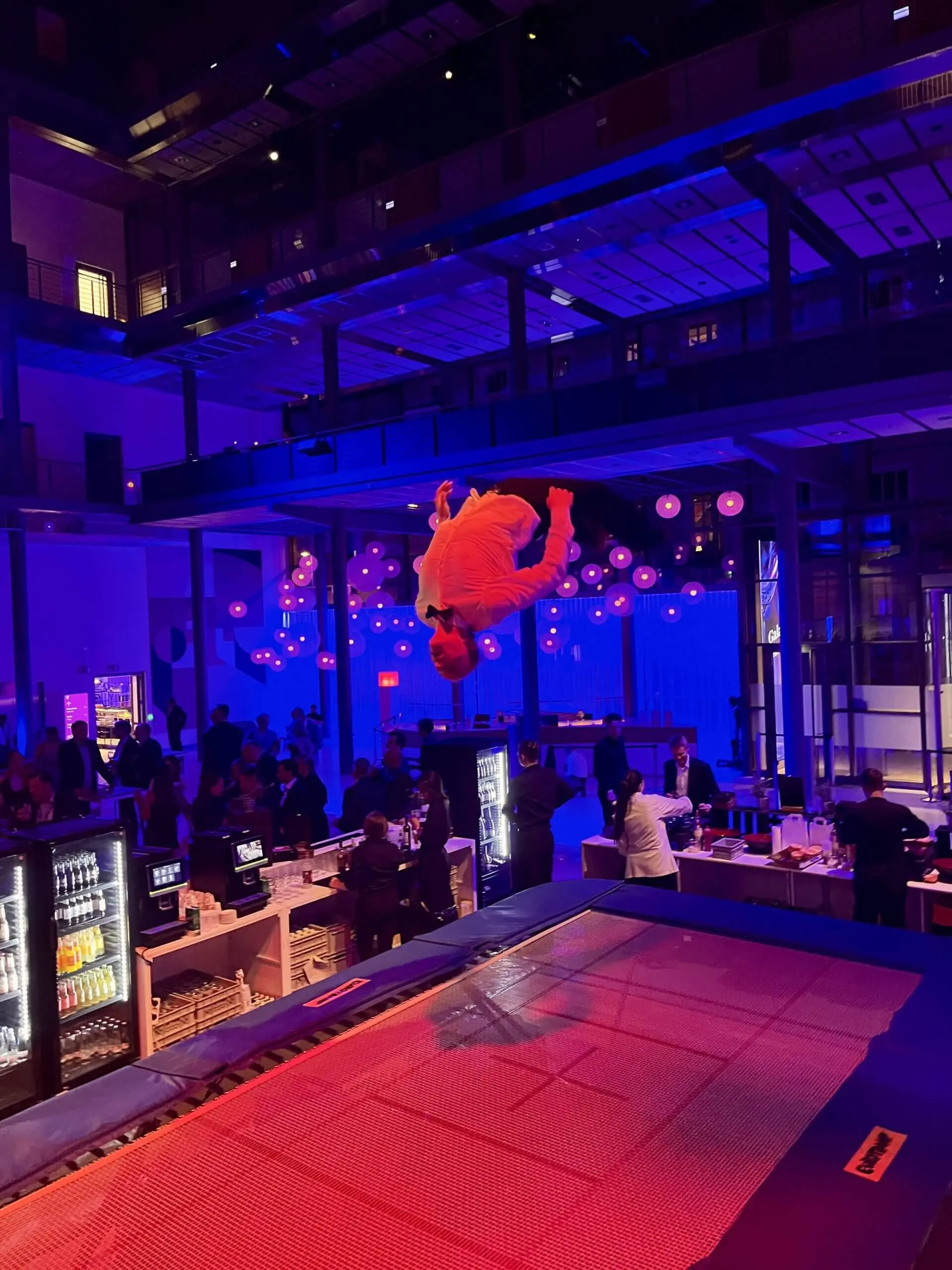 Ein Freestyle-Künstler in einem orangefarbenen Outfit wird mitten in der Luft beim Salto über ein Trampolin in einer modernen, schwach beleuchteten Halle mit blauer und violetter Beleuchtung eingefangen, umgeben von Zuschauern und einem belebten Barbereich.