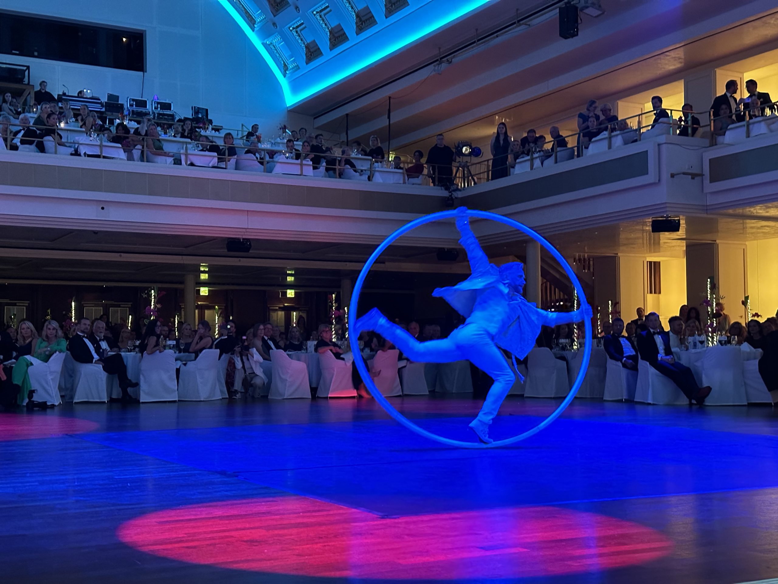 Ein Darsteller im Anzug dreht sich in einem großen Metallreifen auf einer Bühne, die von blauen und rosafarbenen Lichtern beleuchtet wird, während ein elegantes Publikum von Tischen und Balkonen in einem großen Saal aus zusieht.
