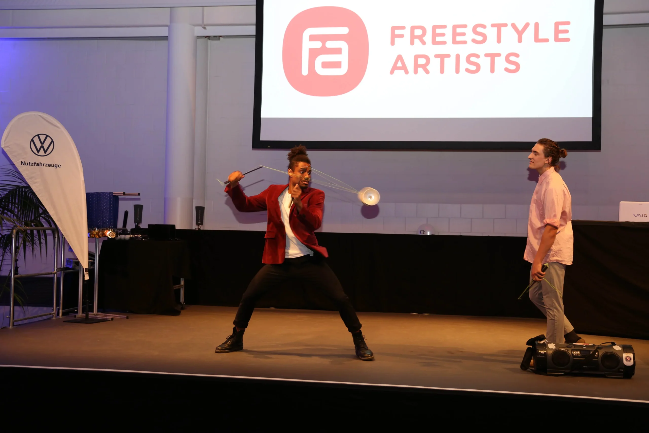 Zwei Künstler stehen auf der Bühne; einer wirbelt gekonnt ein Diabolo als Teil einer Duo-Diabolo-Show, während der andere in der Nähe steht und Jonglierkeulen hält. Auf einer großen Leinwand hinter ihnen steht "FREESTYLE ARTISTS", und es sind Bühnenausrüstung und Banner zu sehen.