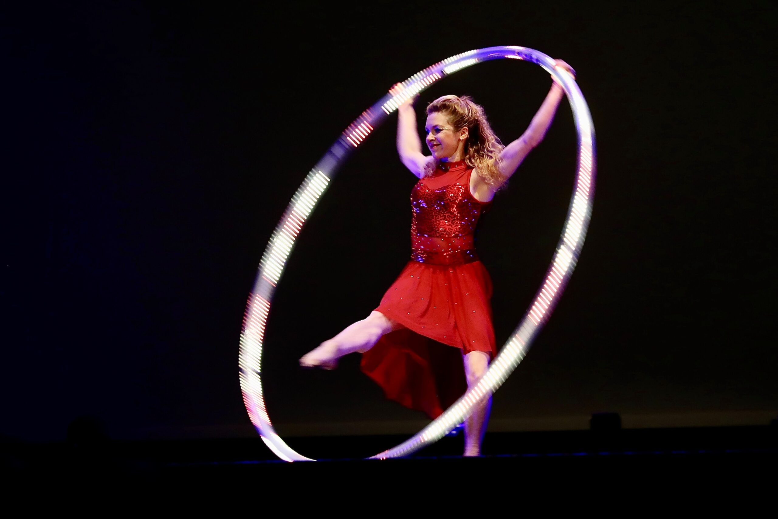 Eine Frau in einem roten Kleid steht auf der Bühne und balanciert in einem großen, beleuchteten Hula-Hoop-Reifen, wobei sie ein Bein hebt und beide Arme den Reifen über ihrem Kopf halten.