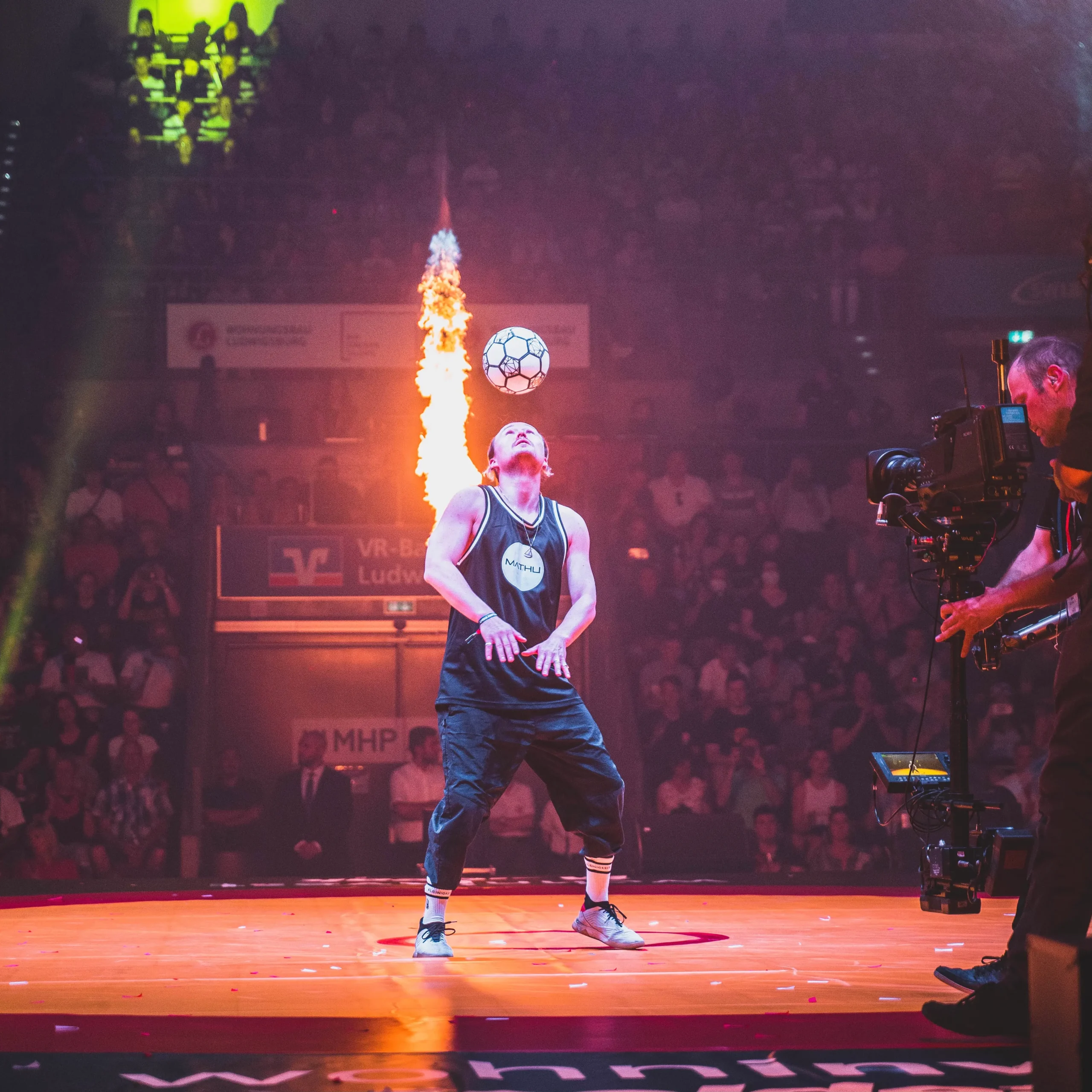 Ein Fußball-Freestyler balanciert auf einer hell erleuchteten Bühne einen Fußball auf der Stirn, während hinter ihm ein Feuerstoß hochschießt und ein Kameramann in der Nähe filmt. Das Publikum schaut von der Tribüne im Hintergrund zu.