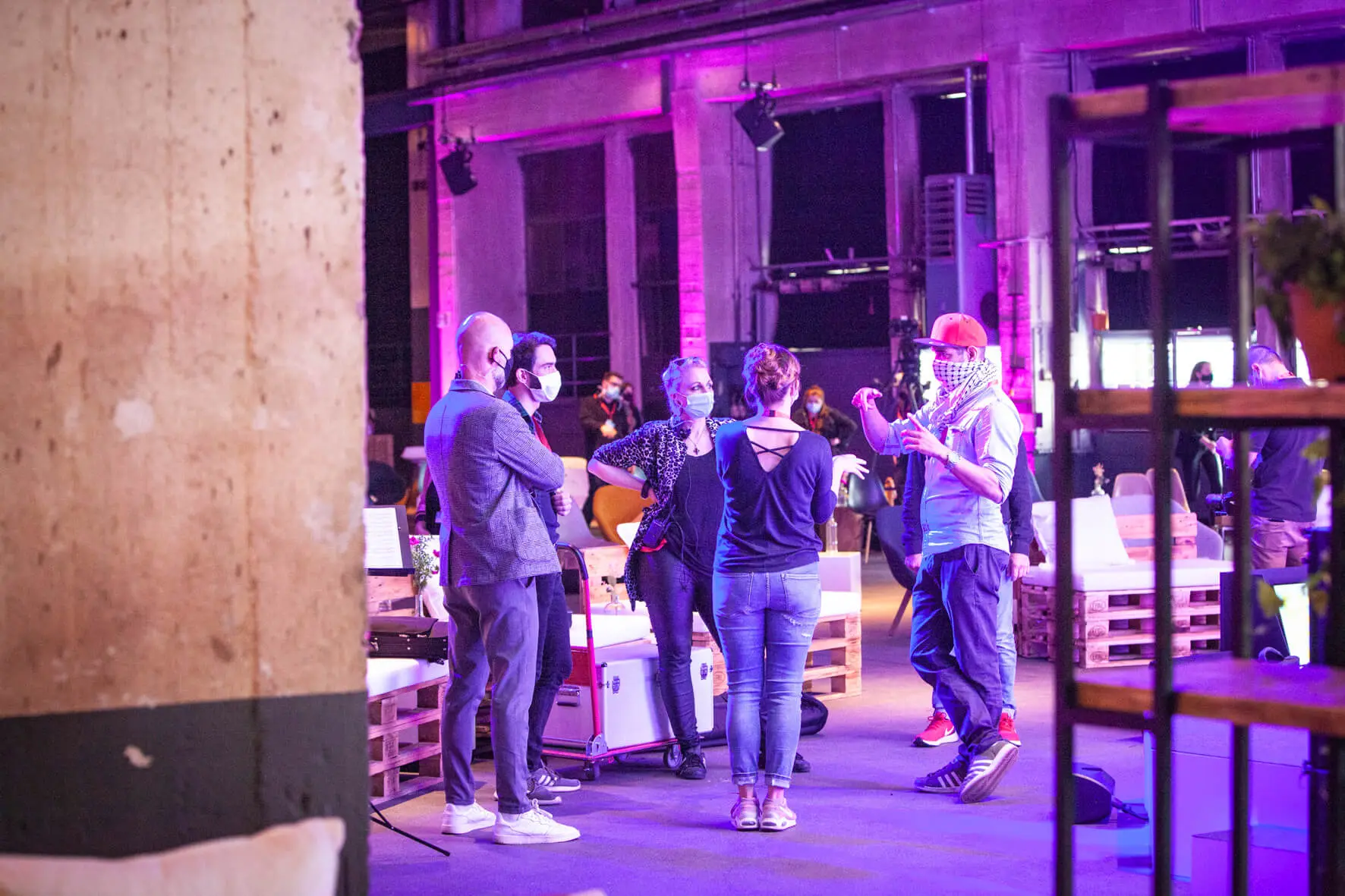 Eine kleine Gruppe von Menschen mit Gesichtsmasken steht im Kreis und unterhält sich in einem geräumigen, modernen, lila beleuchteten Back2Live-Raum mit Sichtbeton und Holzmöbeln.