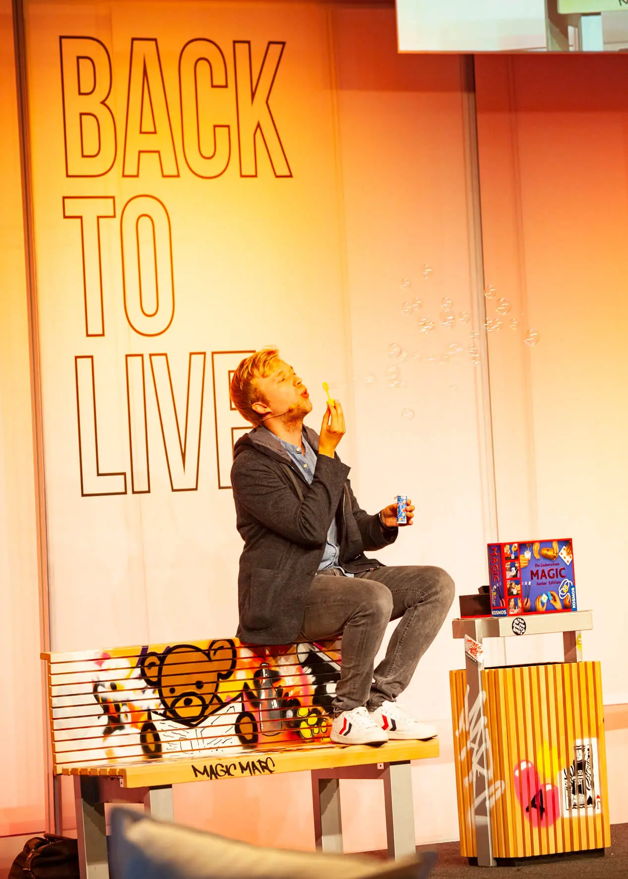 Eine Person sitzt auf einer mit Graffiti bedeckten Bank und pustet Seifenblasen, in der Nähe steht eine Spielzeugkiste. Im Hintergrund steht in großen, fetten Buchstaben "BACK TO LIVE", was den farbenfrohen und spielerischen Geist des Back2Live-Events widerspiegelt.