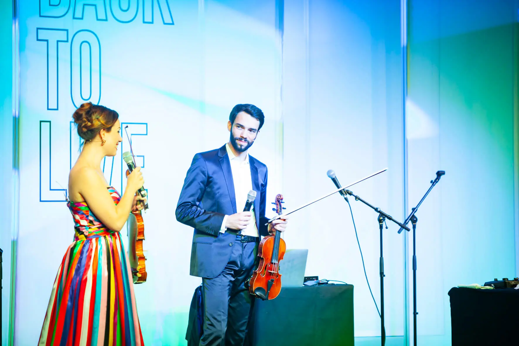 Eine Frau in einem bunt gestreiften Kleid und ein Mann im Anzug, beide mit Geigen in der Hand, stehen auf der Bühne und lächeln sich an. Mikrofone und ein Laptop stehen daneben. BACK TO LIFE und Back2Live stehen an der Wand hinter ihnen.