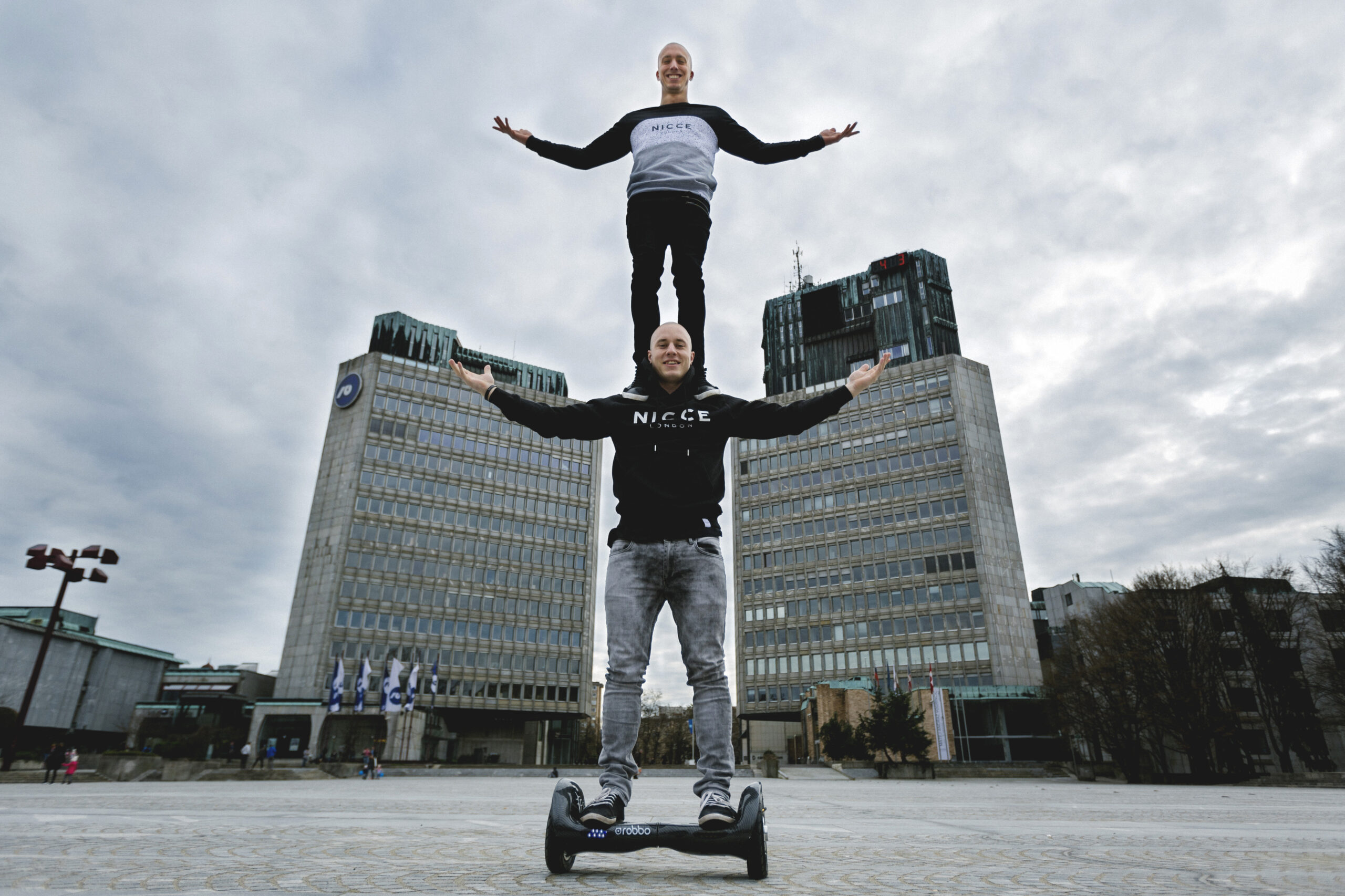 Zwei Männer vollführen einen Balanceakt: Der eine steht mit ausgestreckten Armen auf einem Hoverboard, während der andere auf seinen Schultern steht, ebenfalls mit ausgestreckten Armen, auf einem städtischen Platz mit hohen Gebäuden im Hintergrund.
