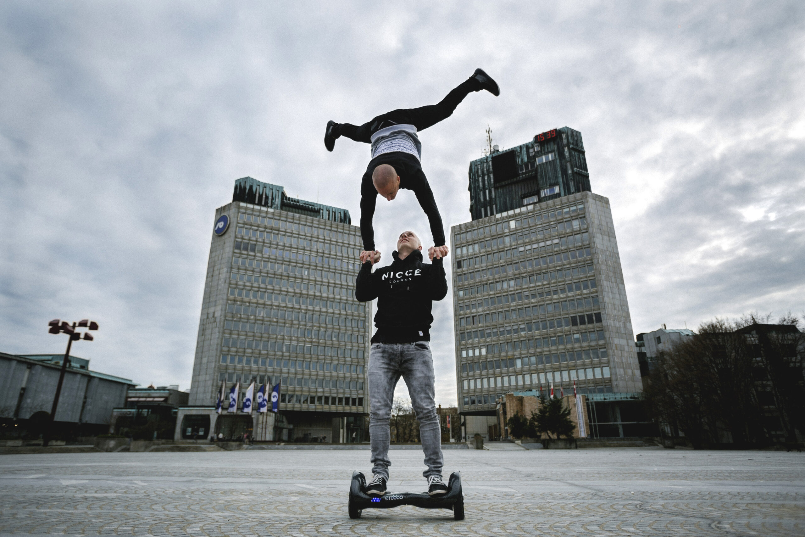 Zwei Personen vollführen im Freien einen akrobatischen Stunt, wobei eine Person auf einem Hoverboard balanciert und die andere kopfüber über ihren Kopf hebt, vor dem Hintergrund moderner Bürogebäude.