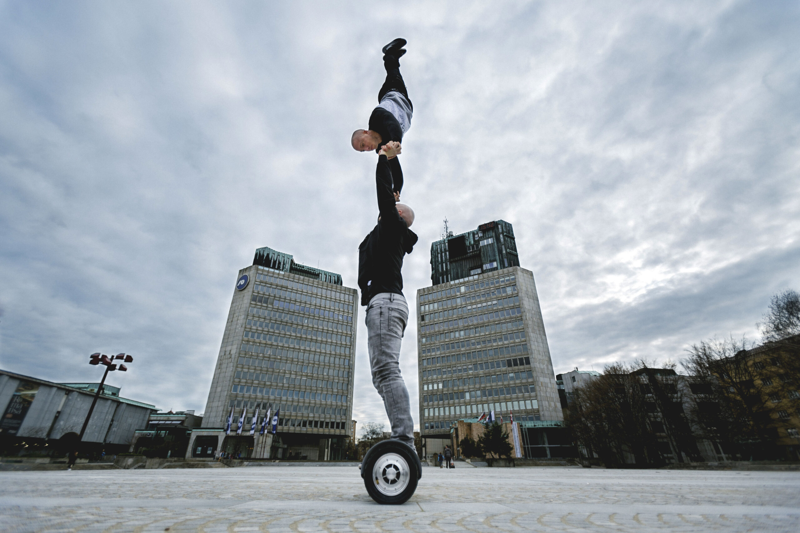 Zwei Personen vollführen auf einem städtischen Platz einen akrobatischen Balanceakt auf einem Einrad, wobei eine Person die andere kopfüber festhält. Im Hintergrund sind hohe Gebäude und ein bewölkter Himmel zu sehen.
