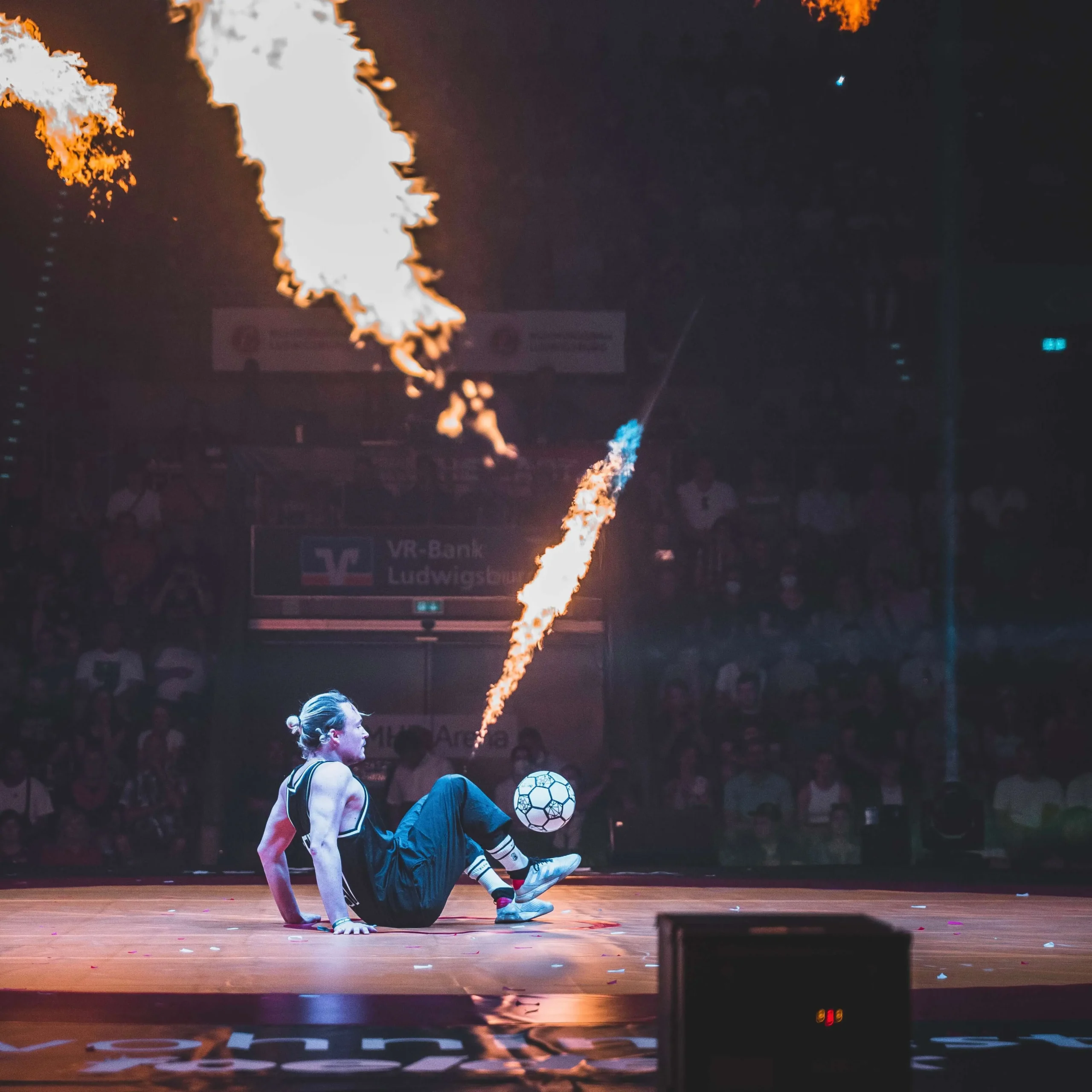 Ein Fußball-Freestyler sitzt im Scheinwerferlicht auf dem Boden und führt Fußballtricks mit einem Ball vor, während Feuerstöße nach oben schießen, während ein Publikum in einer abgedunkelten Arena zusieht.